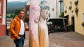 IDEA UNIKA 2025: 6ª edizione della Biennale d’arte nei centri dei paesi della Val Gardena Uomo con giacca arancione accanto a grandi sculture di legno con occhiali da sole in strada pedonale