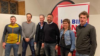 News Sechs Personen stehen vor einem Banner mit rotem und blauem Text in einem Raum