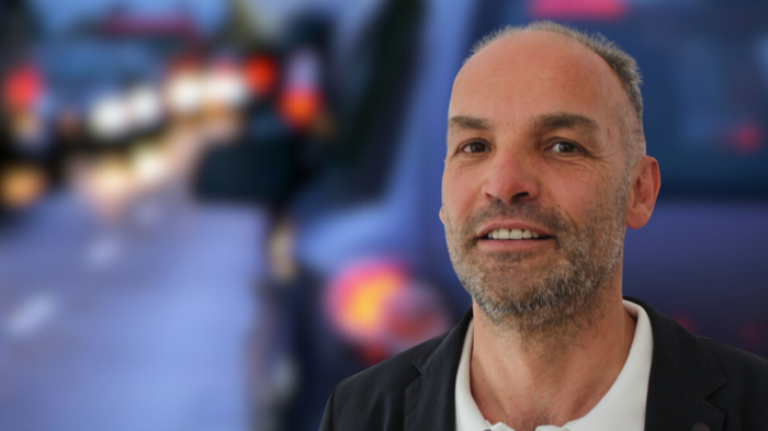 „Megastaus sollten verhindert werden“ Mann mit schwarzem Sakko vor unscharfem Hintergrund mit Fahrzeugen bei Dämmerung