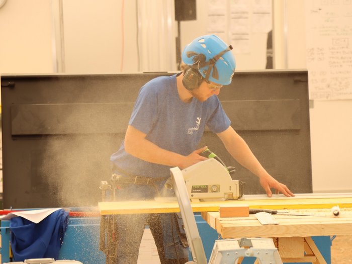 EuroSkills 2021 Mann mit Helm und Schutzbrille sägt Holz mit Kreissäge auf einer Werkbank