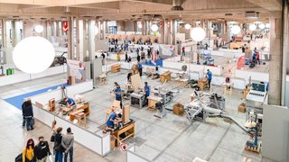 News Große Halle mit Handwerkern bei Bauarbeiten auf Werkbänken und Besuchern