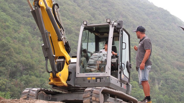 Kinder und Jugendliche lernen die Welt der Baumaschinen Kind lernt Baggerfahren mit Erwachsenem im Freien vor bewaldetem Hügel
