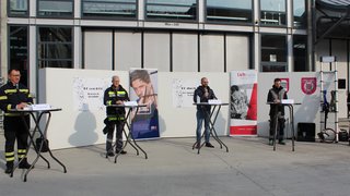News Mehrere Männer stehen an Tischen vor einem Gebäude, einige in Feuerwehruniformen