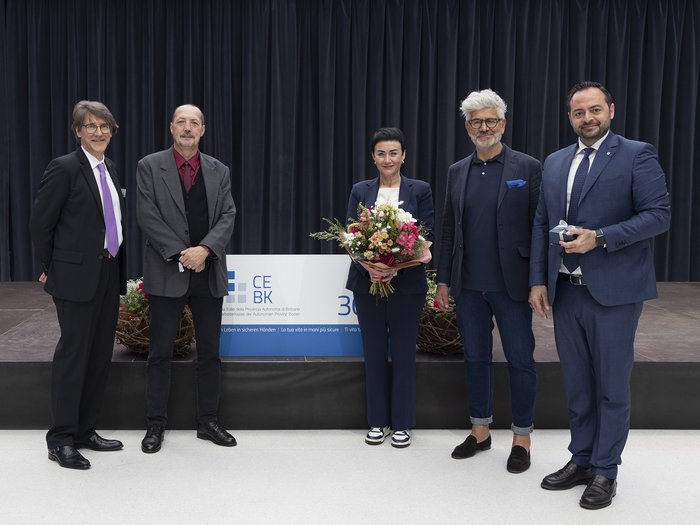 Bauarbeiterkasse der Autonomen Provinz Bozen: Ehrung der Mitglieder „30 Jahre Vertrauen“ Fünf Personen posieren vor einem Vorhang, eine hält Blumen, eine ein Geschenk.