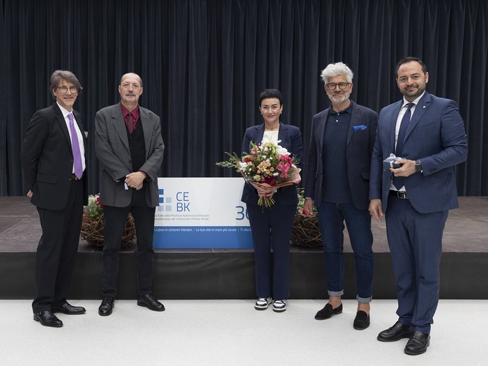 Bauarbeiterkasse der Autonomen Provinz Bozen: Ehrung der Mitglieder „30 Jahre Vertrauen“ Fünf Personen posieren vor einem Vorhang, eine hält Blumen, eine ein Geschenk.