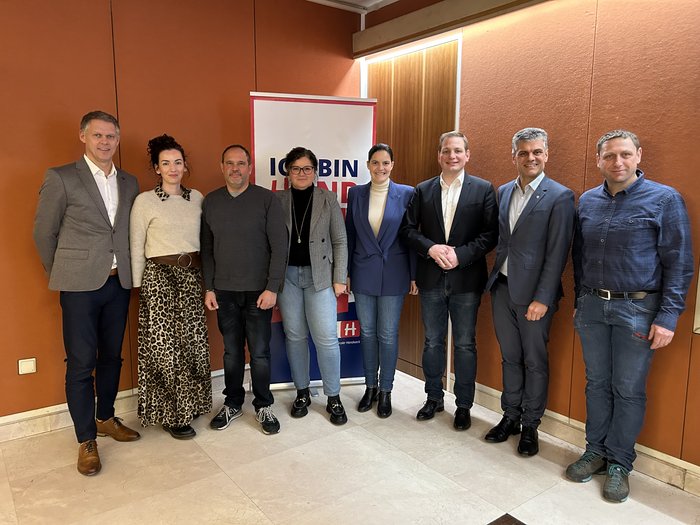 Arbeit durch Bürokratie erschwert Acht Personen stehen zusammen vor einer Werbetafel mit orangefarbenem Hintergrund