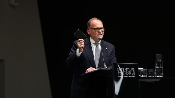 Generalversammlung des SWR 2025 - Ist unsere Wettbewerbsfähigkeit in Gefahr? Mann in Anzug hält ein Gerät und spricht an einem Mikrofon am Rednerpult