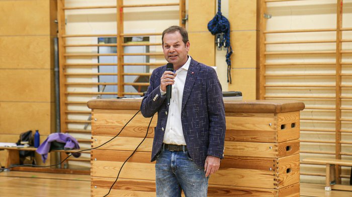Kreative Spielideen: Preisverleihung für "Clever gebaut" Mann spricht mit Mikrofon in einer Turnhalle vor Holzmöbeln