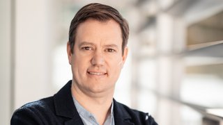 News Porträt eines Mannes mit Hemd und Jacke, der mit verschränkten Armen lächelt