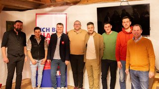 News Acht Männer posieren lächelnd in einem Raum mit Holzdecke und Banner