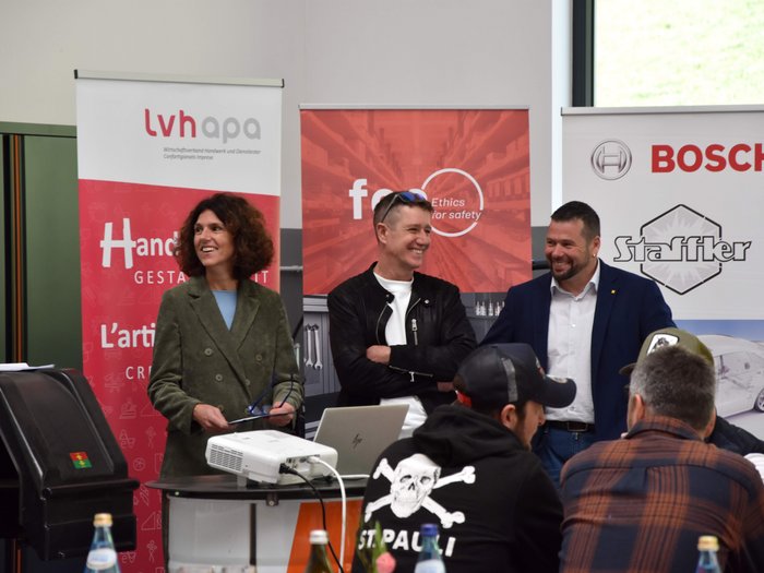 Ein Fokus auf Wettbewerbe und gesetzliche Neuerungen Drei Personen lächelnd vor Werbebannern bei einer Veranstaltung