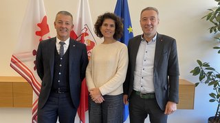 News Drei Personen vor drei Fahnen, lächelnd und formell gekleidet