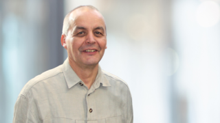 News Uomo con camicia grigia che sorride davanti a sfondo sfocato