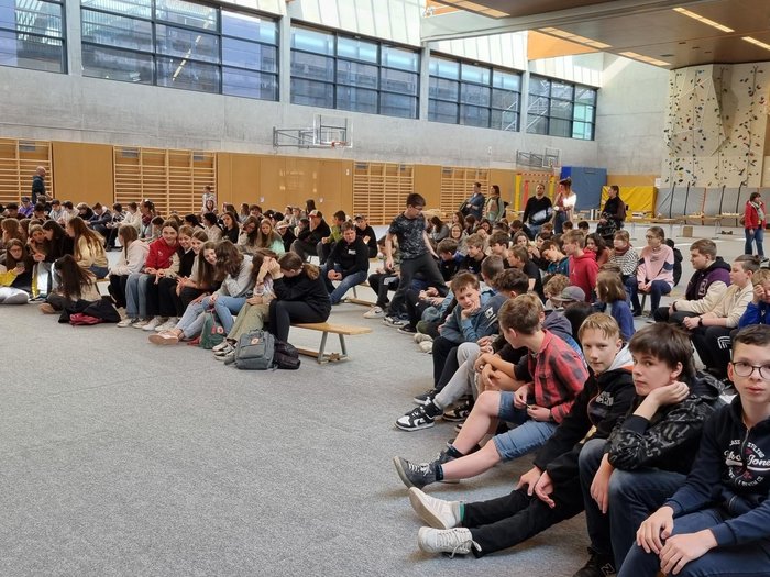 Schulprojekt "Clever gebaut": Selbst entwickelte Spiele prämiert Viele Schüler sitzen in einer Sporthalle auf Bänken und dem Boden