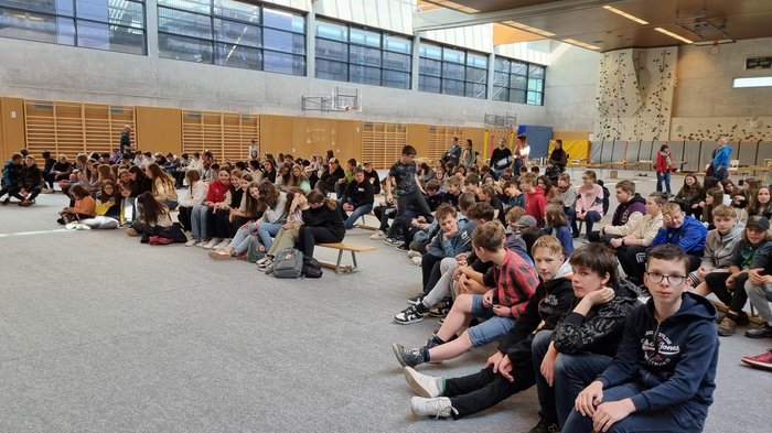 Schulprojekt "Clever gebaut": Selbst entwickelte Spiele prämiert Viele Schüler sitzen in einer Sporthalle auf Bänken und dem Boden