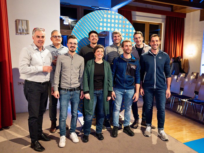 Rittner Musterschau verbindet Menschen und Ideen Neun Personen posieren in einem Raum mit Stühlen und rotem Vorhang für ein Gruppenfoto