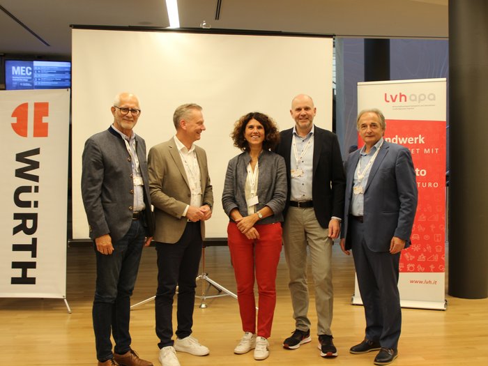 Quattro Paesi s’incontrano a Bolzano per parlare di autovetture: importante dialogo e scambio d’idee Cinque persone in piedi davanti a uno schermo e striscioni pubblicitari a un evento