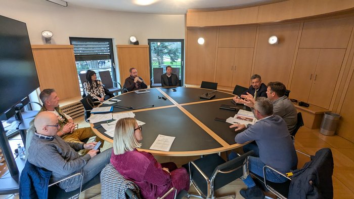 Le imprese movimento terra sostengono l’introduzione di elenchi stradali digitali per una maggiore pianificabilità Nove persone sedute intorno a un tavolo rotondo in una sala riunioni.