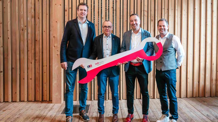 Nominato il nuovo direttore di lvh.apa: Walter Pöhl assume la direzione Quattro uomini in abiti formali tengono una grande chiave inglese rossa davanti a una parete di legno