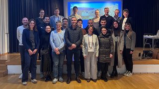 News Gruppenfoto von 21 Erwachsenen in einem Veranstaltungsraum vor einer Projektionsleinwand