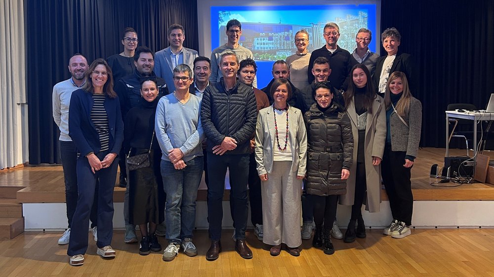 Home Gruppenfoto von 21 Erwachsenen in einem Veranstaltungsraum vor einer Projektionsleinwand