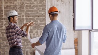 News Zwei Bauarbeiter mit Helmen besprechen Pläne in einem Rohbau mit offener Wand