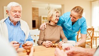 News Pflegekraft schaut lächelnd ältere Menschen beim Spielen eines Brettspiels an