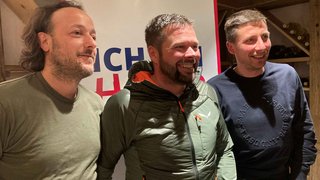 News Drei Männer lachen vor einem Banner bei einer Veranstaltung