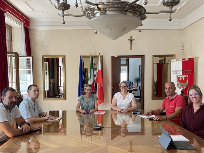 Lieferverkehr in Meran: Lösungen für ein verkehrsberuhigtes Zentrum Sechs Personen sitzen an einem großen Holztisch in einem Raum mit Flaggen und Kreuz an der Wand