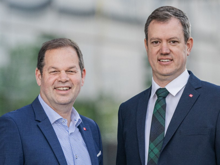 “Wir stehen zur Verfügung” Zwei Männer in Anzügen lächeln vor unscharfem Hintergrund