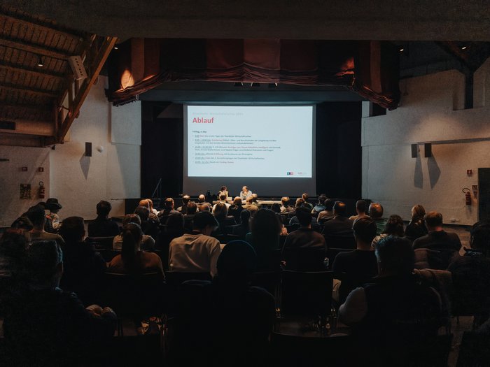 Ritorna la Fiera dell‘Economia Val d’Isarco: una vetrina della diversità Pubblico ascolta una conferenza durante una mostra economica in una sala buia