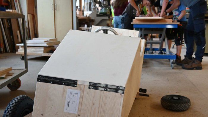 Seifenkistenrennen in St. Felix: Handwerk im Rausch der Geschwindigkeit Holzseifenkiste im Vordergrund in Werkstatt mit Jugendlichen im Hintergrund