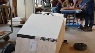 News Holzseifenkiste im Vordergrund in Werkstatt mit Jugendlichen im Hintergrund