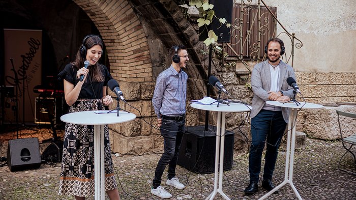 Generation-H Aperitif Drei Personen mit Kopfhörern sprechen an Stehtischen in einem Innenhof