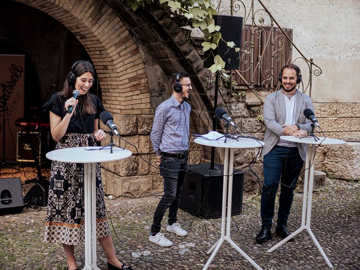 Generation-H Aperitif Drei Personen mit Kopfhörern sprechen an Stehtischen in einem Innenhof