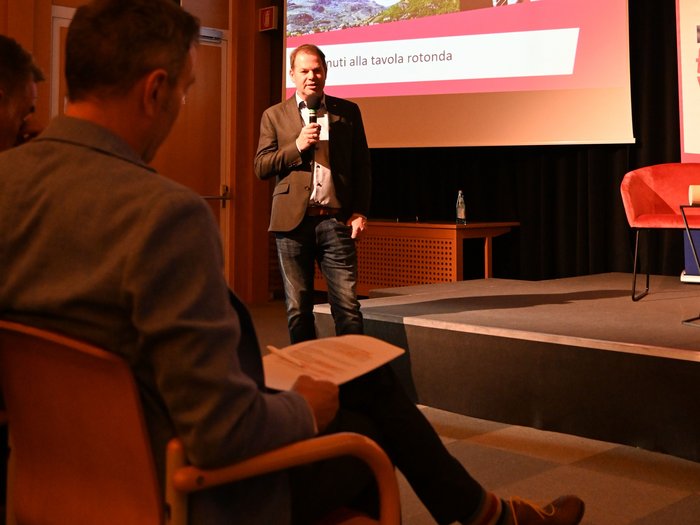 Gesprächsrunde „Die Stadt, die du dir wünschst“ – Ein Blick auf die Zukunft von Bozen Mann spricht mit Mikrofon vor Publikum in Konferenzraum mit Präsentation