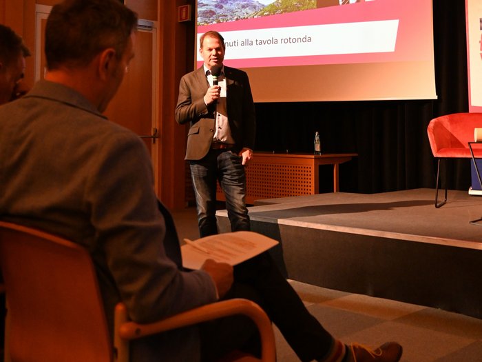 "La città che desideri": un incontro per il futuro di Bolzano Uomo parla con microfono davanti al pubblico in sala conferenze con presentazione