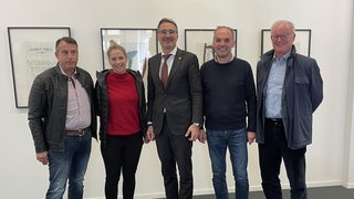 News Fünf Personen stehen nebeneinander vor einer weißen Wand mit gerahmten Bildern