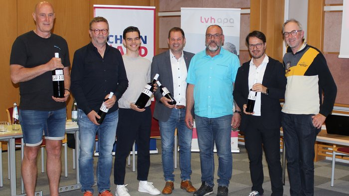 20 Jahre Erfolg und Sicherheit Sieben Männer stehen in einem Raum, vier halten Weinflaschen in der Hand