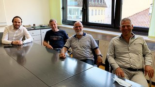 News Vier Männer sitzen um einen Konferenztisch in einem Büro mit Fenster im Hintergrund.