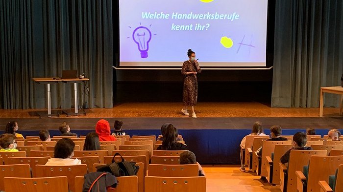 Junghandwerker und HGJ in Mals und Glurns zu Besuch Lehrerin mit Maske spricht vor Kindern in einem Hörsaal über Handwerksberufe