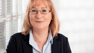 News Porträt einer Frau mit Brille, Bluse und Blazer vor heller Glaswand