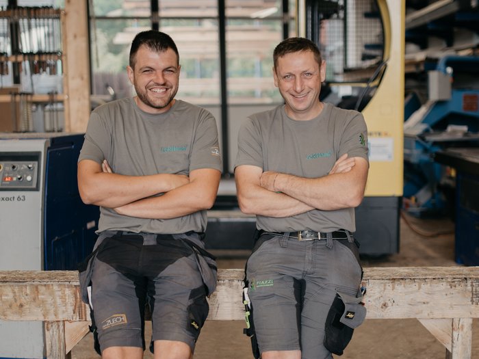 Zwei erfahrene Zimmermänner initiieren Crowdfunding-Kampagne für nachhaltiges Bauen Zwei lächelnde Handwerker sitzen in Werkstatt und tragen graue T-Shirts