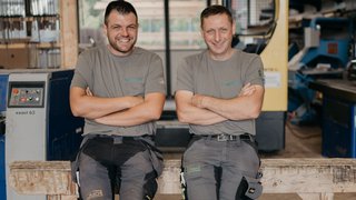 News Zwei lächelnde Handwerker sitzen in Werkstatt und tragen graue T-Shirts
