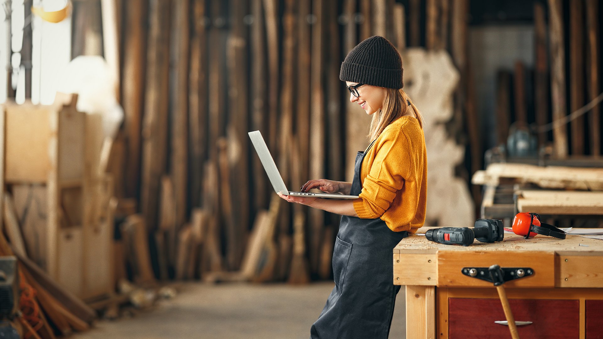 Home Donna con berretto in bottega usa laptop vicino a legno e attrezzi