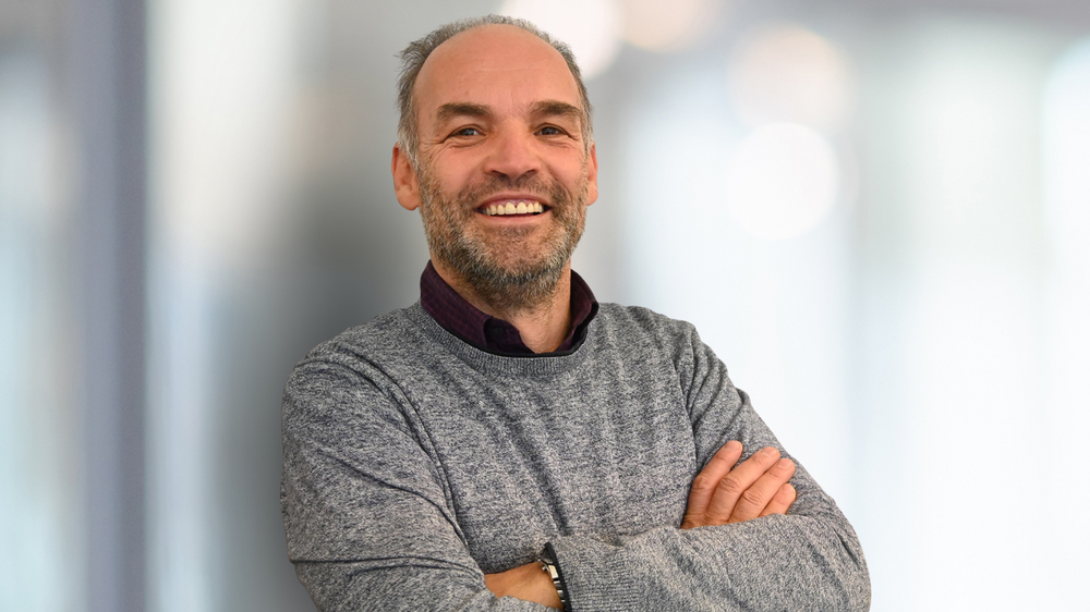 Uomo sorridente con maglione grigio e braccia incrociate su sfondo chiaro