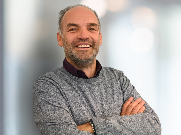 „Kein Interesse an gelenkter Lieferverzögerung“ Lächelnder Mann mit grauem Pullover und verschränkten Armen vor hellem Hintergrund