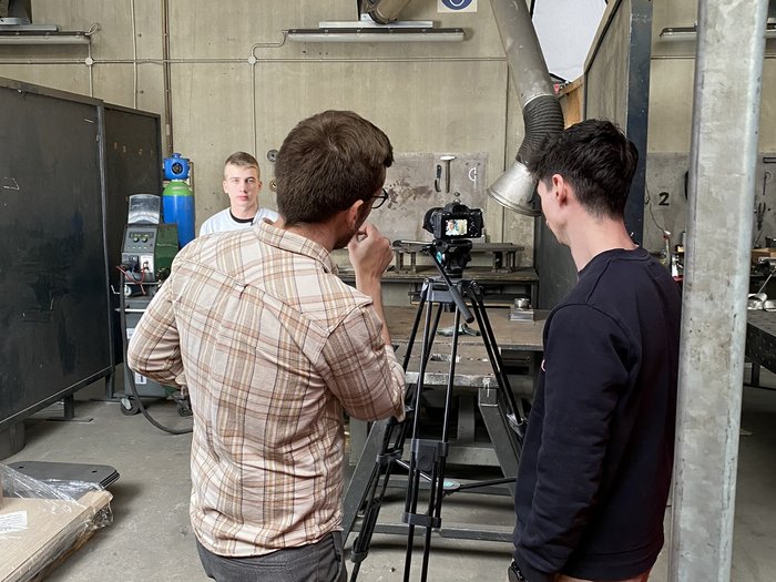„Was du baust, bewegt die Welt“ Zwei Männer filmen in einer Werkstatt, eine Person sitzt vor der Kamera
