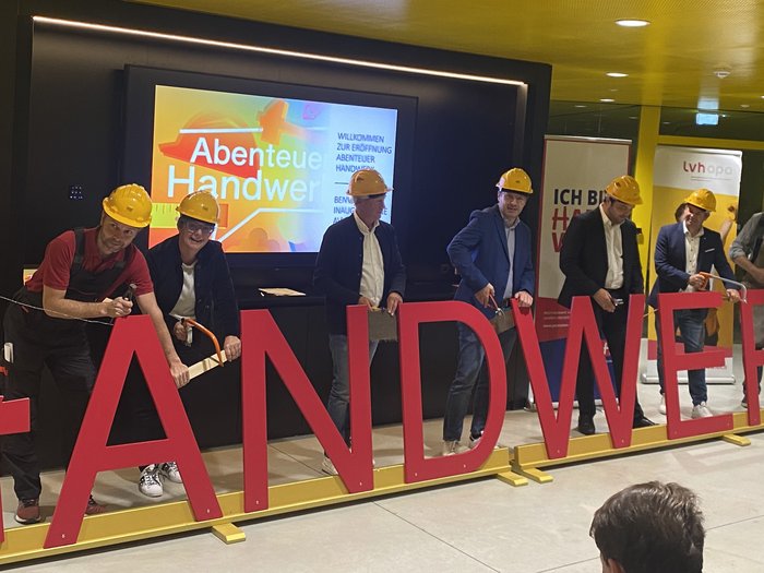 "Abenteuer Handwerk" - Initiative begeistert junge Generation für Handwerksberufe im Pustertal Personen mit Schutzhelmen sägen große rote Buchstaben des Wortes Handwerk