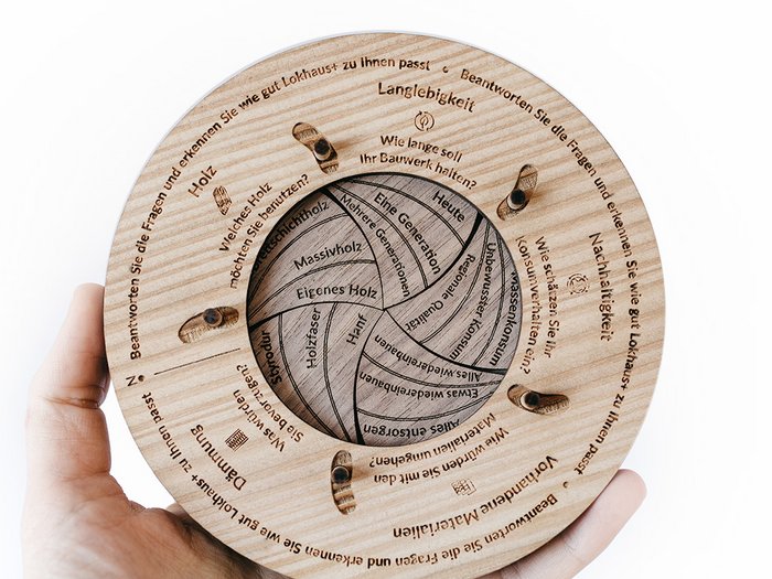 Gleich drei Auszeichnungen in Platin Hand hält runden, beschrifteten Holzdiagramm mit Fragen zu Holz und Nachhaltigkeit